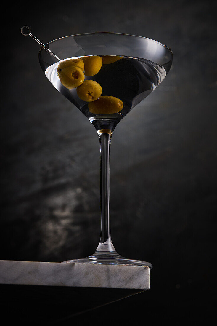 Mit Alkohol gefülltes Glas Martini mit reifen Oliven am Spieß auf Marmortischkante vor dunklem Hintergrund im Studio