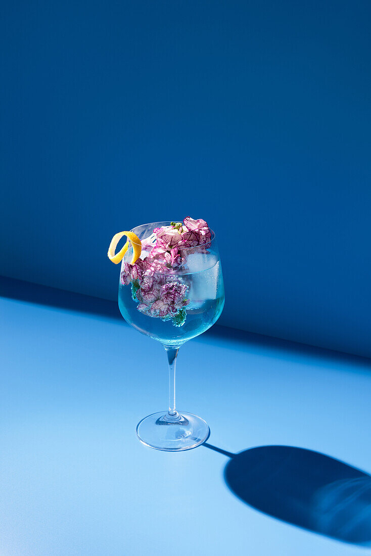 Transparentes Glas mit frischem Cocktail, Minzblättern und Blumen vor blauem Hintergrund auf dem Tisch