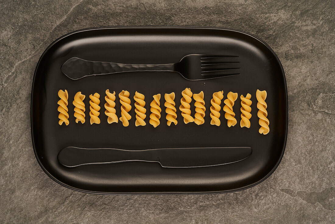 Top view of black fork and knife placed near uncooked fusilli pasta on tray on table