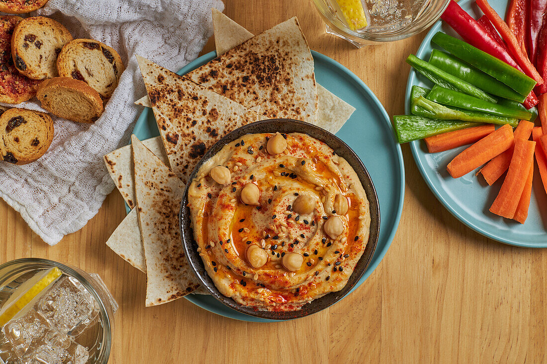 Draufsicht auf köstlichen Hummus und Tortilla-Chips auf einem Teller neben verschiedenen Gemüsesorten und Croutons und Gläsern mit kalter Limonade auf einem Holztisch