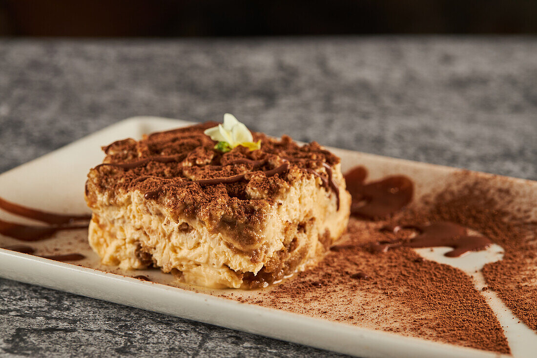 Piece of delicious tiramisu dessert garnished with cocoa powder and chocolate sauce with flower and served on rectangular plate with berries on gray marble table in restaurant