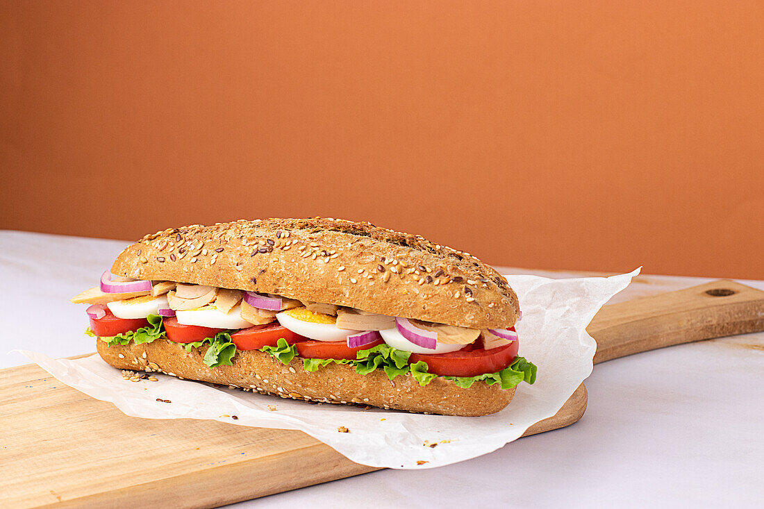 Appetizing sandwich with hard boiled egg, fresh lettuce, tuna and tomatoes served on baking paper on chopping wooden board on colorful background