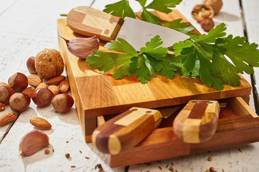 Walnüsse mit Mandeln und Haselnüssen auf einem hölzernen Schneidebrett mit Petersilie und Knoblauch neben Messern auf einem weißen Holztisch