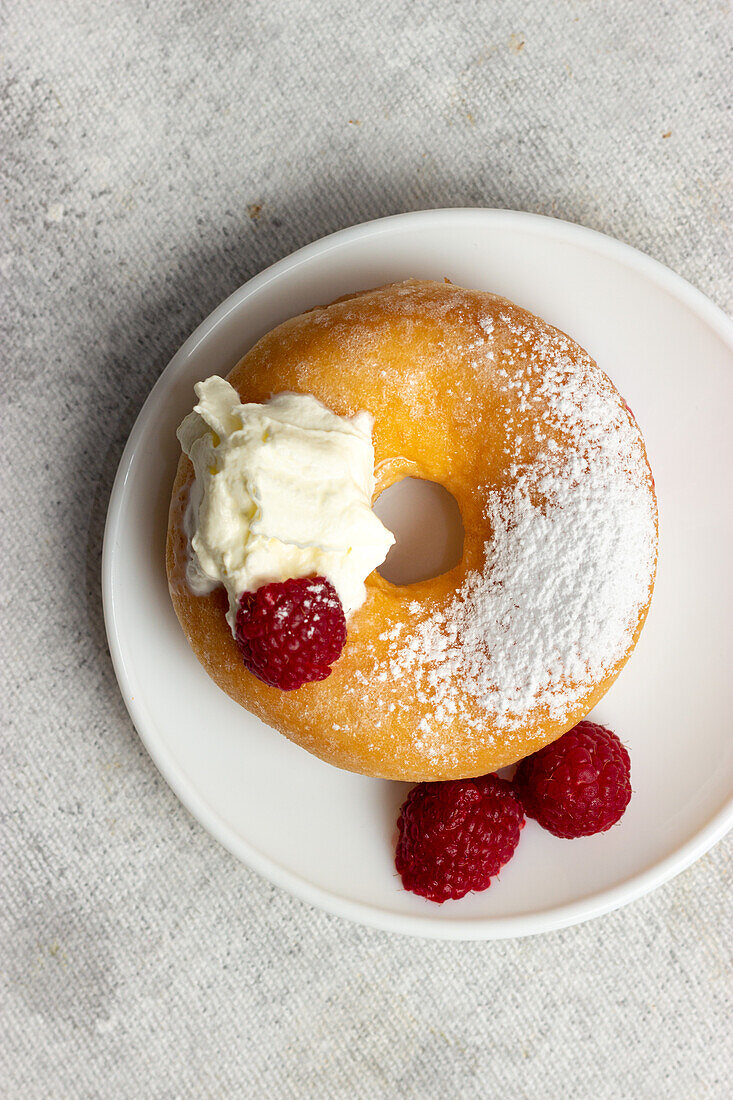 Top view of tasty doughnut sprinkled with powdered sugar served on plate with fresh raspberries on gray background