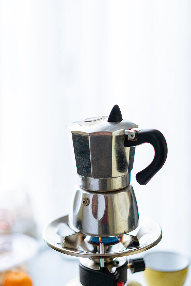 Moderne Metall-Moka-Kanne zum Aufbrühen von Kaffee auf einem rostfreien Gasherd in einer hellen Küche mit Tassen im unscharfen Hintergrund