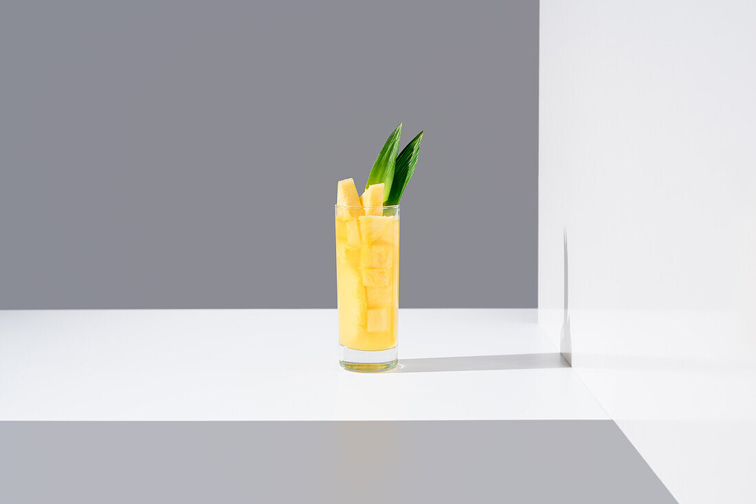 Glass full of cold pineapple juice with green leaves and straw placed on sunlit table against gray and white background