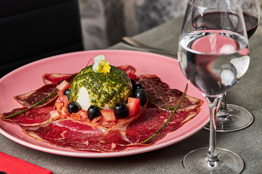 Von oben Tomatensalat mit Burrata und Prosciutto-Scheiben, garniert mit schwarzen Oliven und Pesto-Sauce und serviert auf Teller auf Holztisch