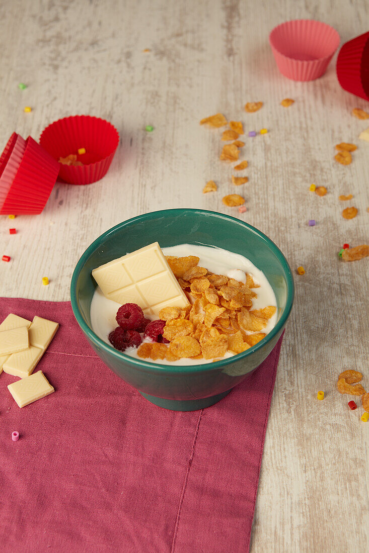 From above bowl of crunchy cornflakes and yogurt garnished with fresh raspberries and white chocolate placed on wooden table near red cupcake cases during breakfast