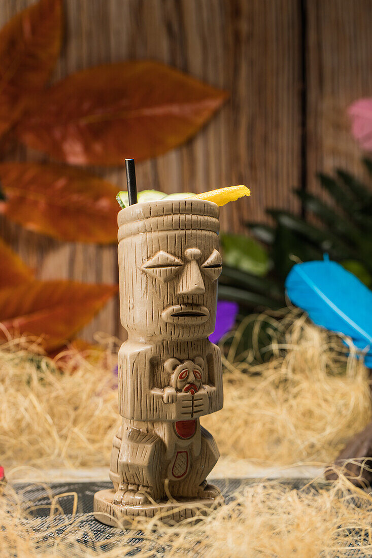Traditioneller skulpturaler Tiki-Becher mit alkoholischem Getränk und Strohhalm auf Teppich vor Holzzaun mit bunten Blättern und trockenem Gras