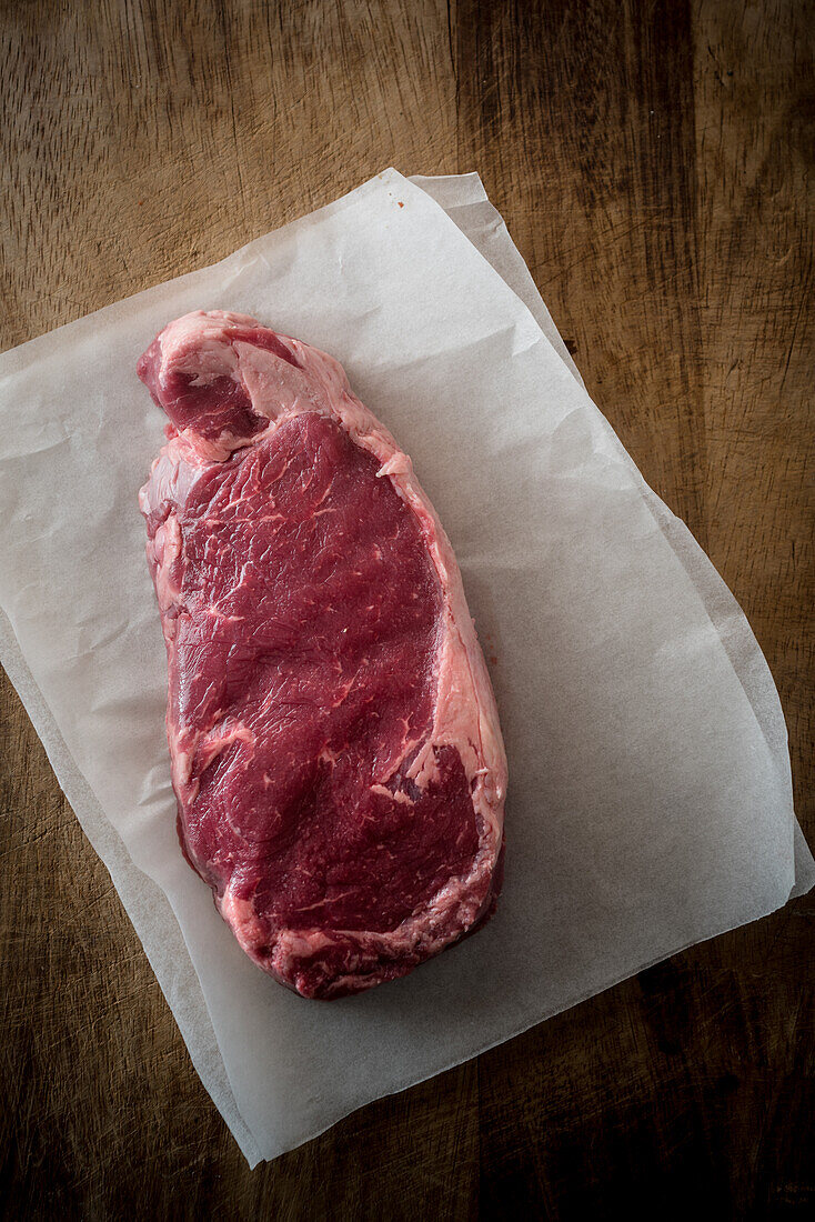 Overhead view of uncooked meat piece with rosemary leaves against baking paper on brown background