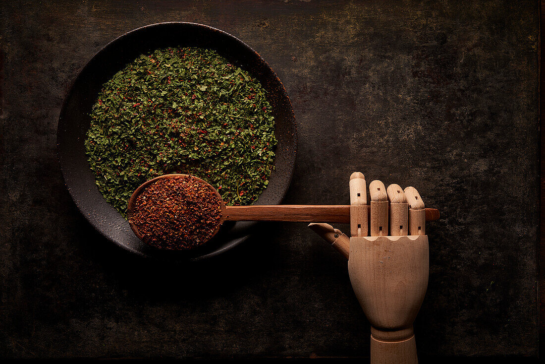 Top view of bowl with green dried herbs and spoon with ground sun dried tomato powder with artificial wooden hand on black background