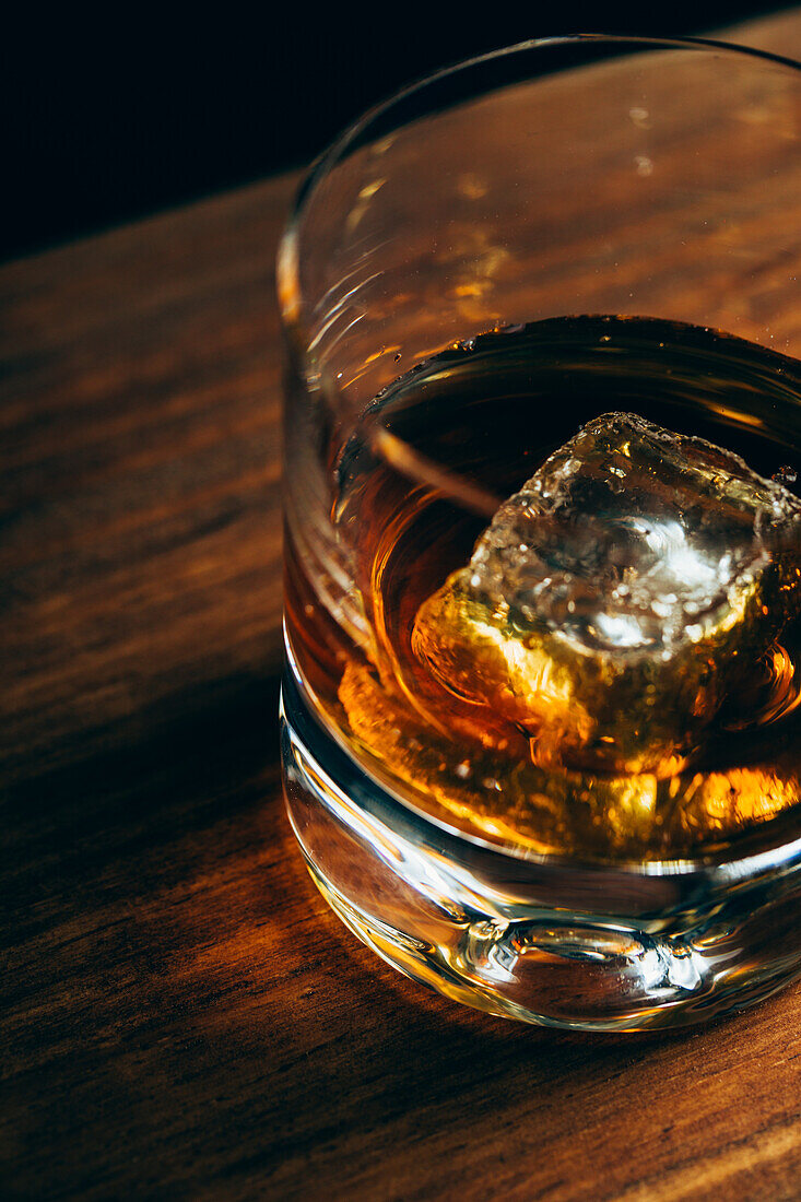 From above glass cup with cold whiskey and cube of ice placed on lumber table in dark room