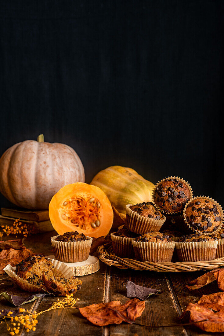 Komposition aus appetitlichen Muffins mit Schokoladenstückchen auf Holztisch zwischen verstreuten Herbstblättern und Kürbissen auf dunklem Hintergrund