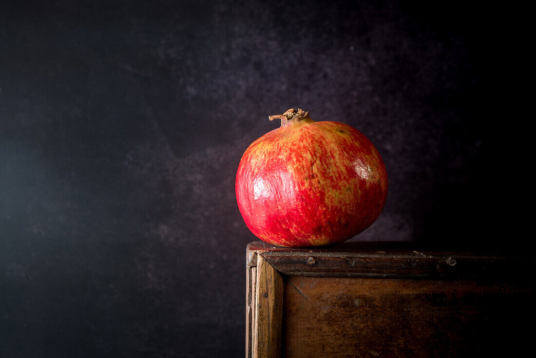 Still life composition with bright red whole pomegranate fruit placed on wooden stand against black background