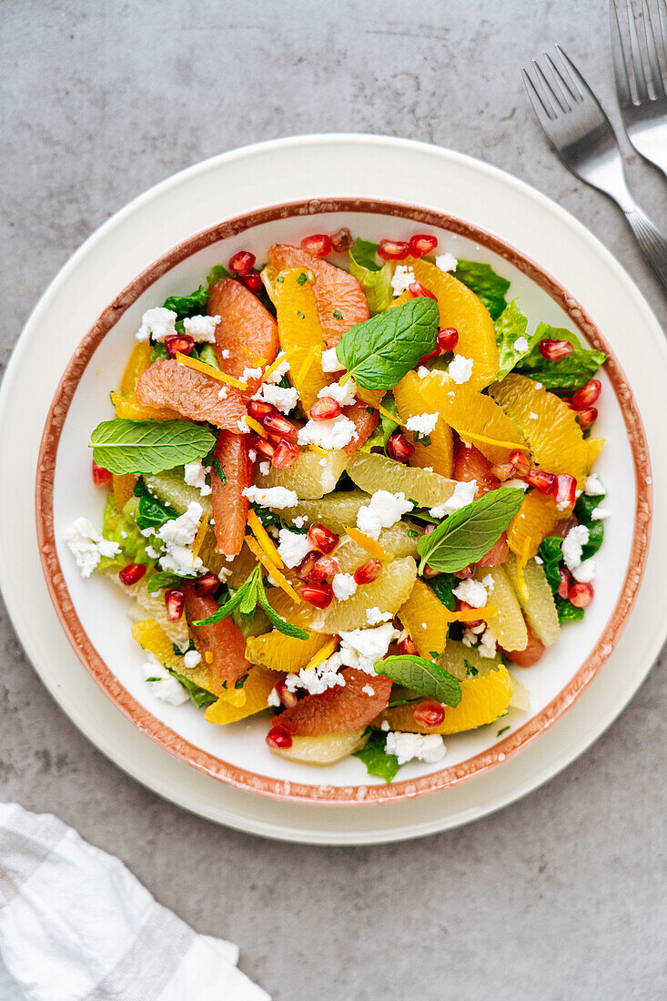 Draufsicht auf traditionellen griechischen Salat mit reifen Tomaten und Feta-Käse, gekrönt von Granatapfelkernen und Minzblättern auf grauem Hintergrund mit Orangensaft