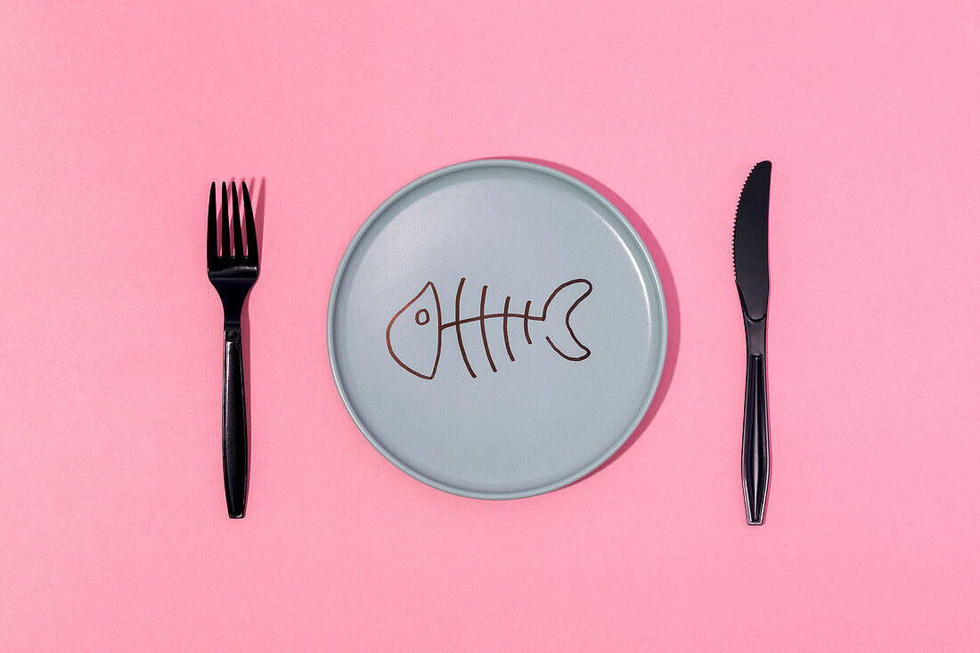 From above of gray ceramic plate with painted fish skeleton served on pink surface with black fork and knife