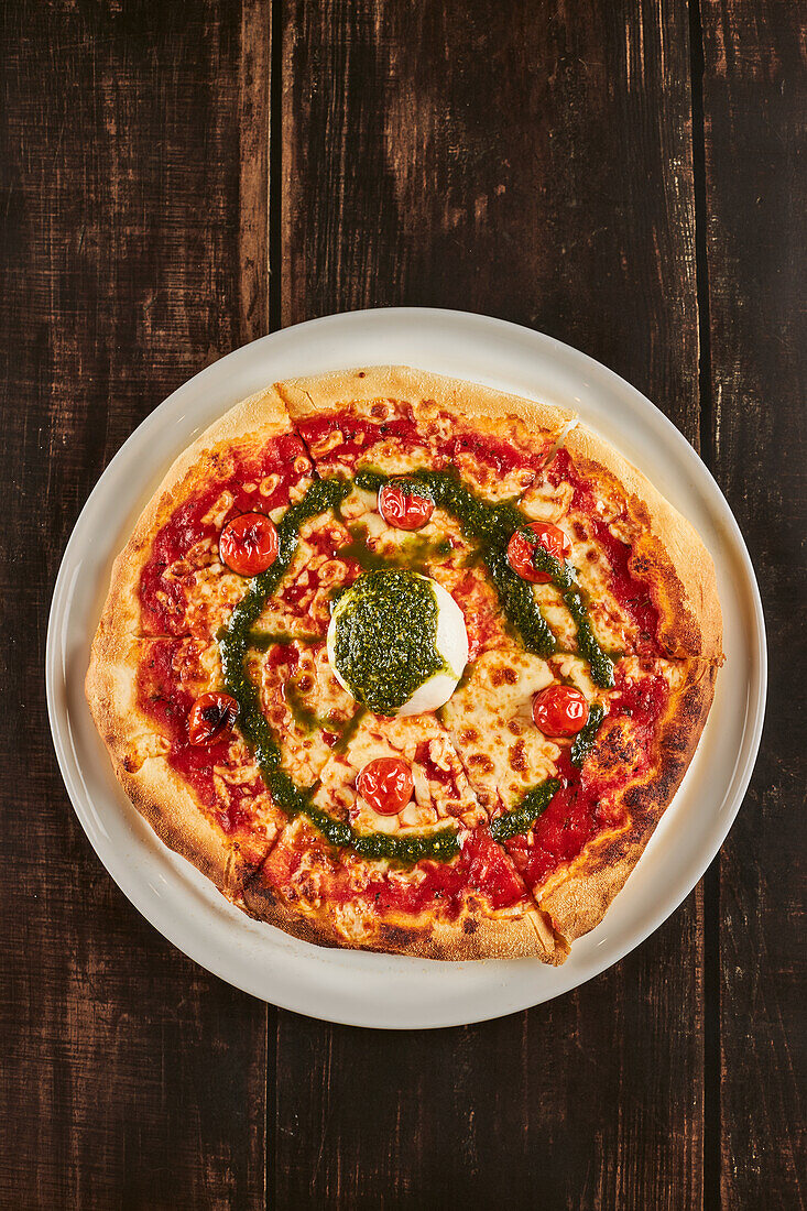 From above delicious pizza with cherry tomatoes and parmesan garnished with mozzarella cheese ball and pesto sauce and served on plate on lumber table in pizzeria