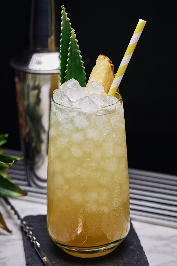 Gelber Cocktail im Glas garniert mit Ananasstück und grünen Blättern mit Papierstrohhalm auf Schieferuntersetzer mit Barlöffel