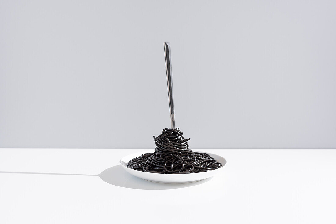 Stainless fork in full bowl of black spaghetti with cuttlefish ink on white table in studio on gray background