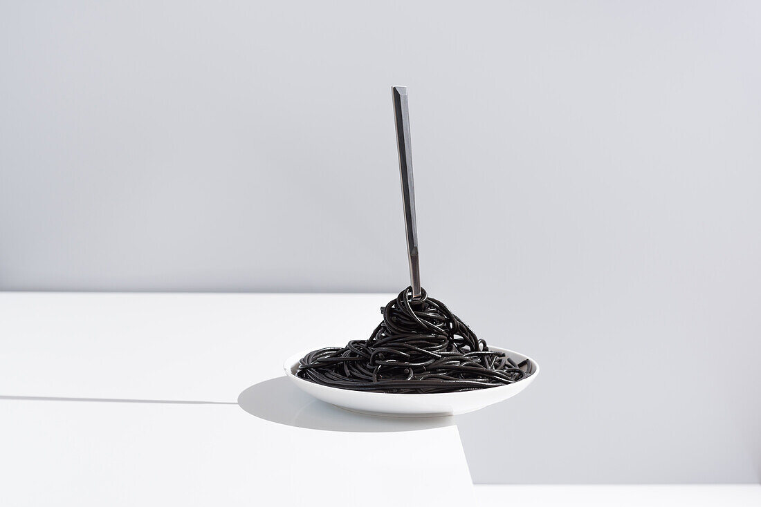 Rostfreie Gabel in voller Schüssel mit schwarzen Spaghetti mit Tintenfischtinte auf weißem Tisch im Studio auf grauem Hintergrund