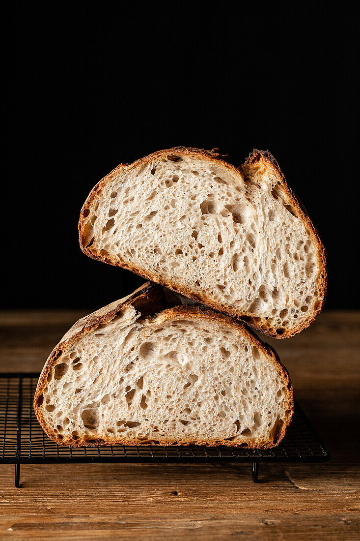 Hälften von schmackhaftem Sauerteigbrot mit brauner Kruste auf einem Grillrost auf einem Holztisch vor schwarzem Hintergrund