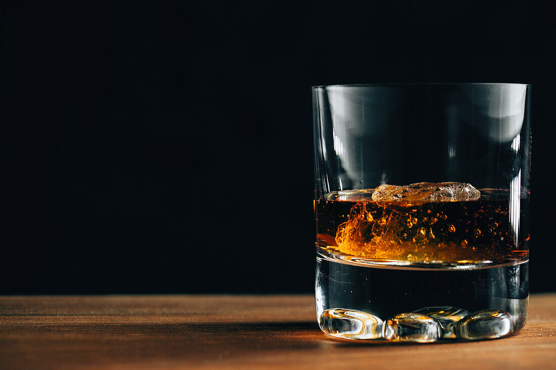 Glass cup with cold whiskey and cube of ice placed on lumber table in dark room