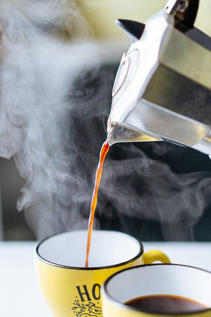 Freshly brewed steaming hot coffee pouring from metal moka pot into yellow cup on table in light kitchen on blurred background