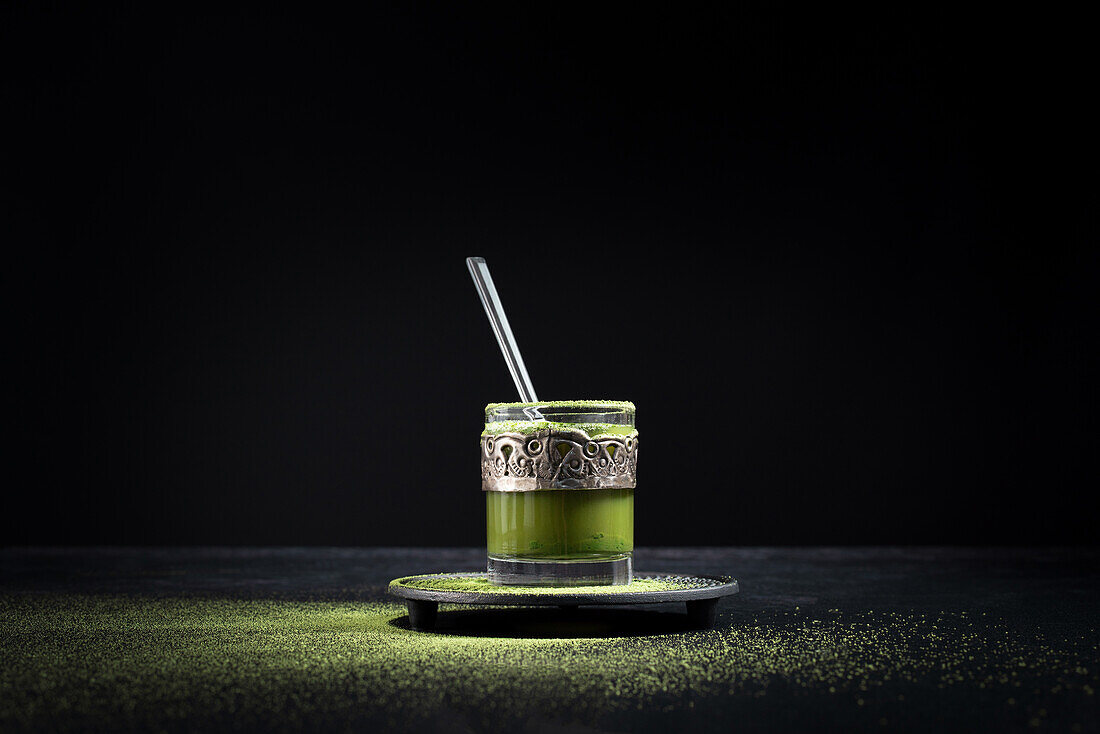 Healthy herbal green matcha tea served in glass cup with metal decoration on saucer sprinkled with powder on black table