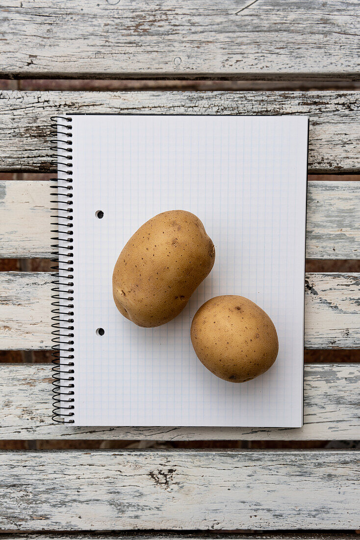 Draufsicht auf zwei Kartoffeln, die auf einem aufgeschlagenen Notizblock mit leeren Seiten auf einem Holztisch liegen