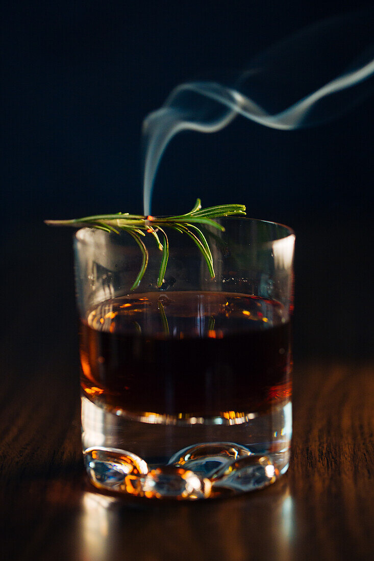 Glass of whiskey with rosemary placed on wooden table against blue background