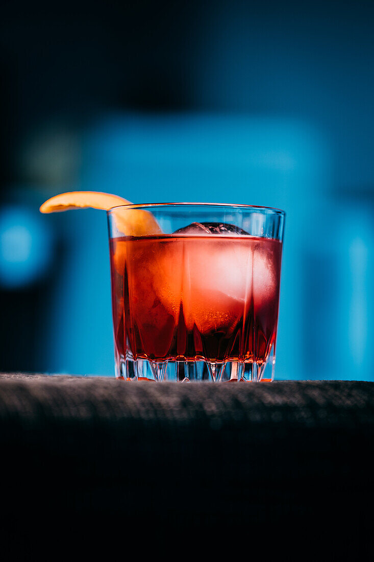 Glas erfrischender Negroni-Cocktail mit bitterem Geschmack und Eis, garniert mit Orangenschalen, serviert auf dem Arm der Couch in einem dunklen Raum