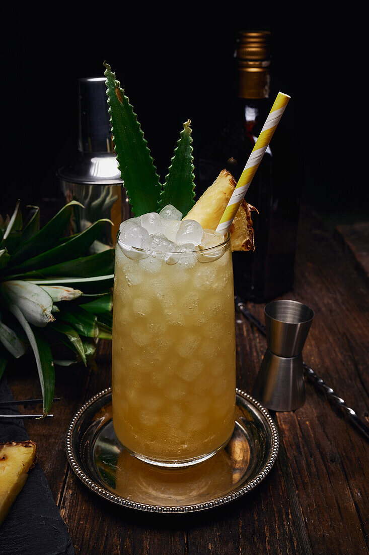 Glass of alcoholic cocktail decorated with pineapple piece and leaves with paper straw on tray near shaker and bottle with shot glass at table on black background