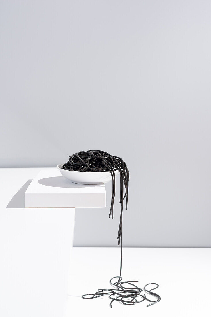 Minimalist studio with black squid ink spaghetti falling out from full ceramic bowl on white table