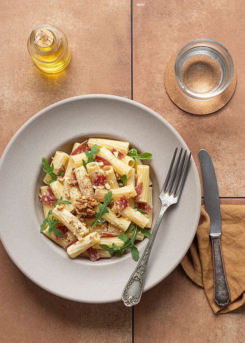 Nudelgericht mit Rucola, Salami und Walnüssen von oben auf einem Tisch, umgeben von Öl, Zitrone und Besteck