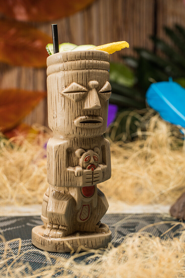 Traditioneller skulpturaler Tiki-Becher mit alkoholischem Getränk und Strohhalm auf Teppich vor Holzzaun mit bunten Blättern und trockenem Gras