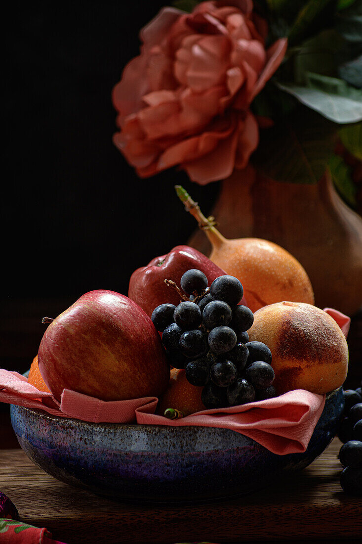 Frische Äpfel und Pfirsiche mit Weintrauben und Granadilla in einer Schale auf einem Tisch neben einer roten Pfingstrose