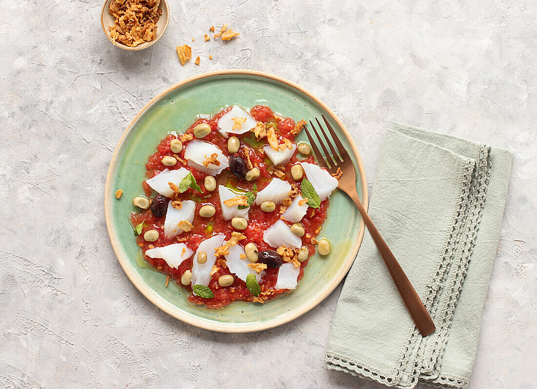 Summery tomato and cod salad in a shallow dish with a fork and a bowl of fried onions