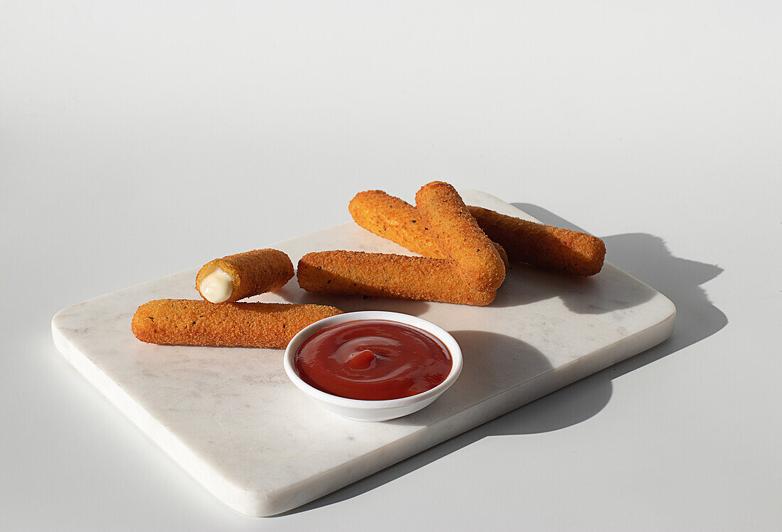 Gebratene Mozzarella-Käse-Sticks in einer Metallfritteuse in der Nähe von Ketchup-Sauce auf einer Marmorplatte auf weißem Hintergrund