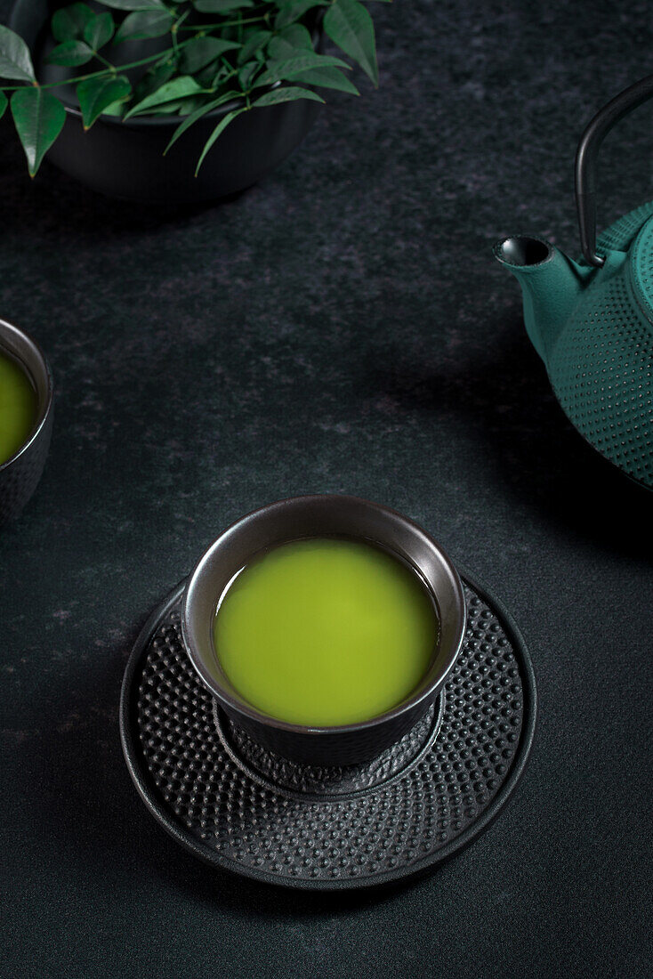 Schwarze Keramiktasse mit traditionellem japanischen grün gefärbten Matcha-Tee, serviert auf einem Tisch mit Teekanne