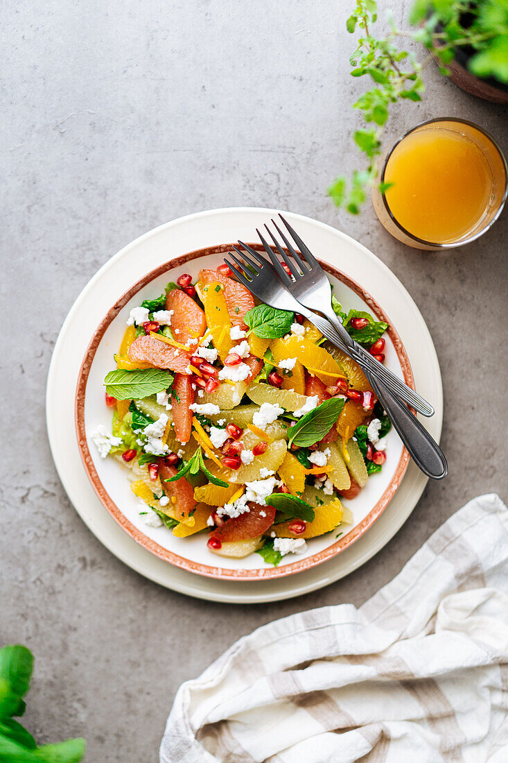 Draufsicht auf traditionellen griechischen Salat mit reifen Tomaten und Feta-Käse, gekrönt von Granatapfelkernen und Minzblättern auf grauem Hintergrund mit Orangensaft