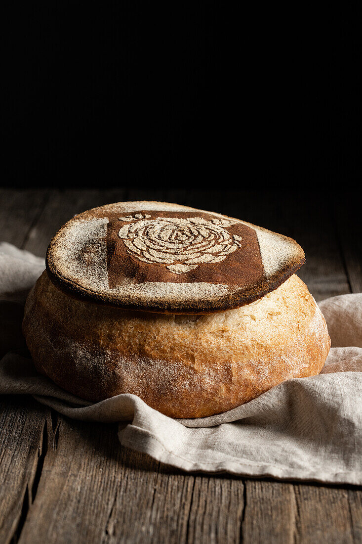 Dekoratives, frisch gebackenes Brot mit Blumenmuster auf einer Serviette auf einem Holztisch in der Küche vor schwarzem Hintergrund
