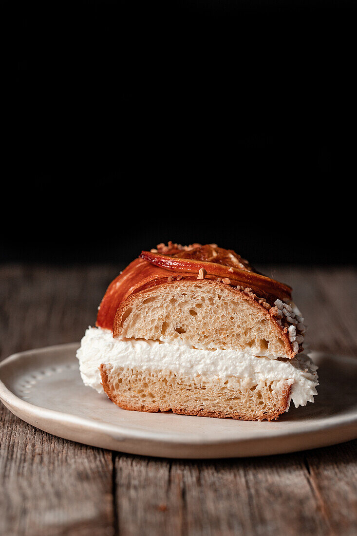 Appetizing slice of king's cake stuffed with white cream on plate placed on wooden table against black background