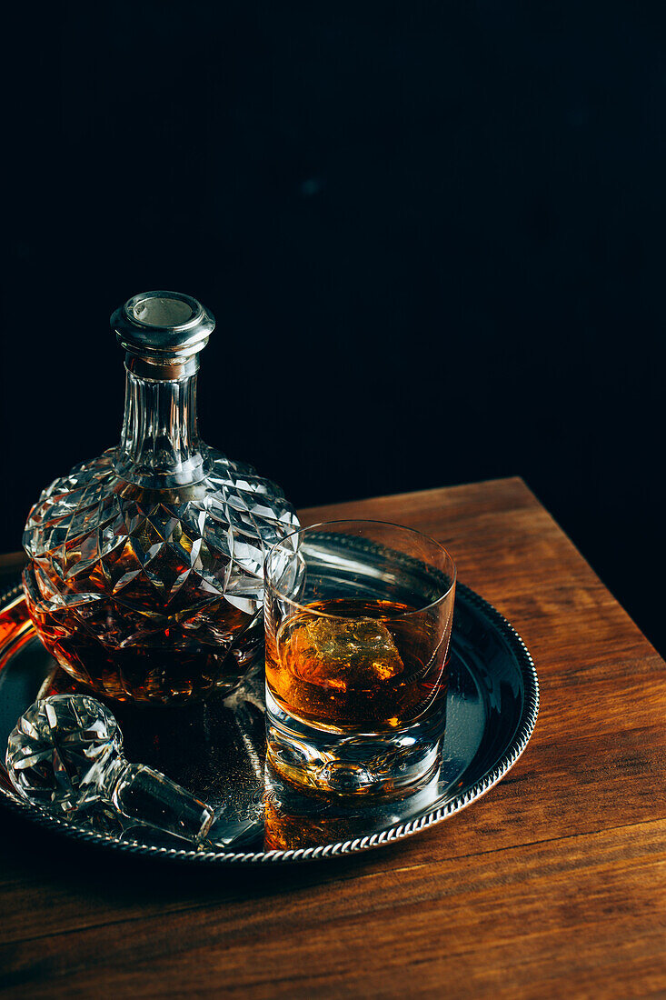 Glass of cold whiskey with ice placed on wooden table near decanter on black background