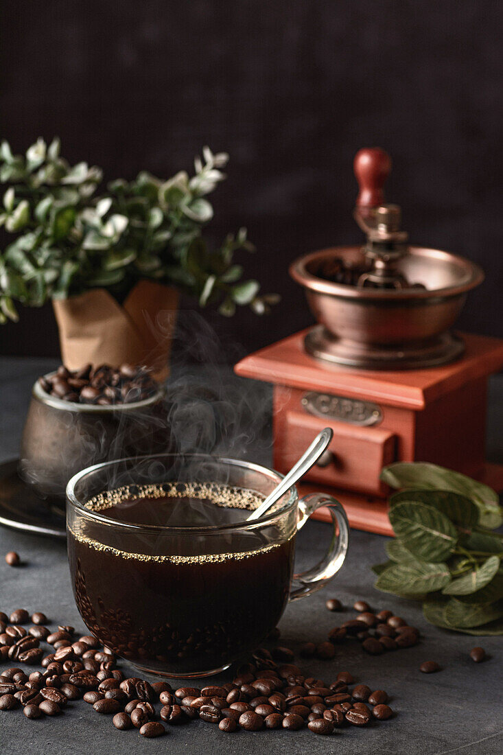 Glass of freshly brewed aromatic coffee on saucer placed on table with scattered beans against manual grinder