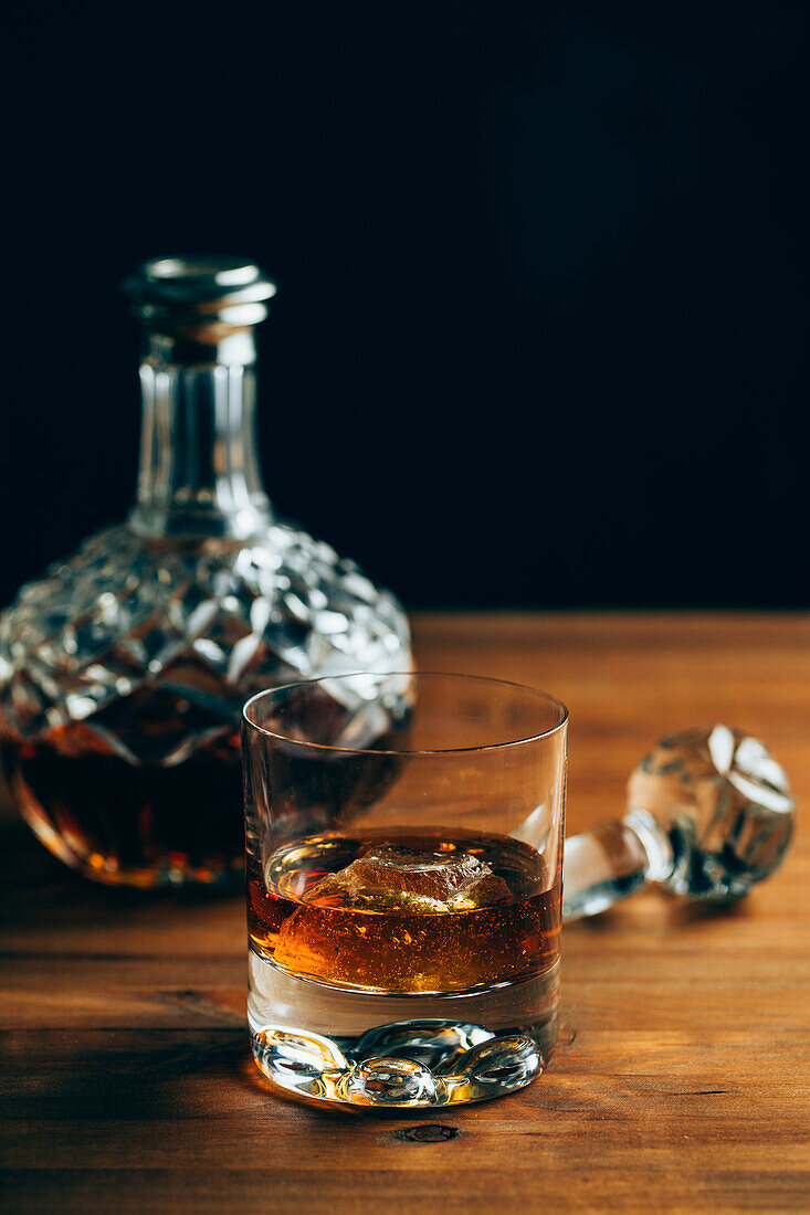 Glass of cold whiskey with ice placed on wooden table near decanter on black background