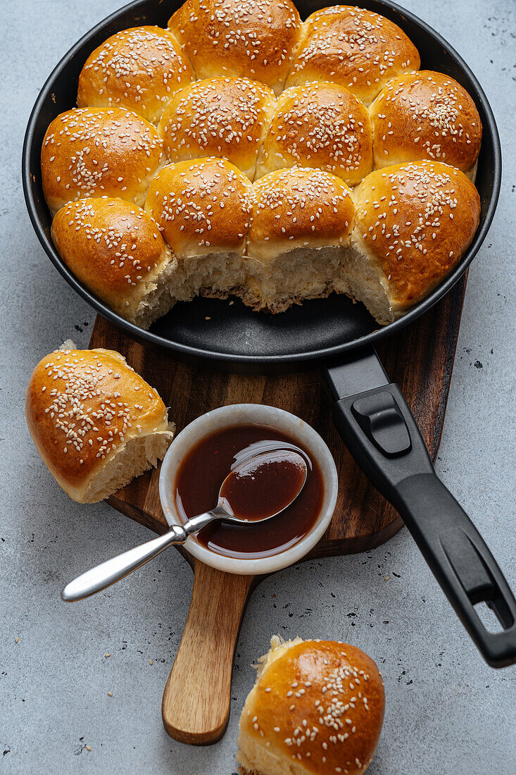 From above of tasty pull apart bread rolls with sesame seeds in frying pan served on wooden cutting board with sweet sauce