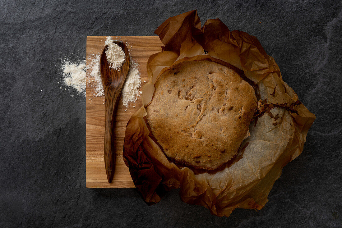 Top view composition with freshly baked rustic sourdough round bread loaf on parchment paper placed on wooden board with spoon and wheat flour