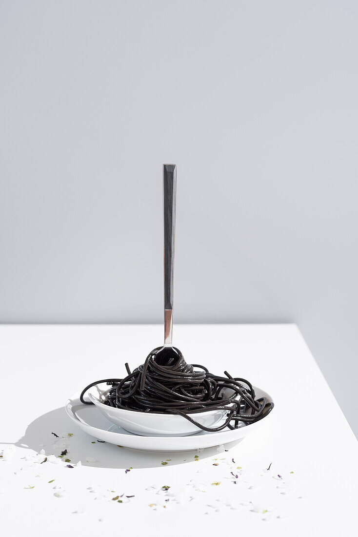 Rostfreie Gabel in voller Schüssel mit schwarzen Spaghetti mit Tintenfischtinte auf weißem Tisch mit dünnen Blüten im Atelier auf grauem Hintergrund