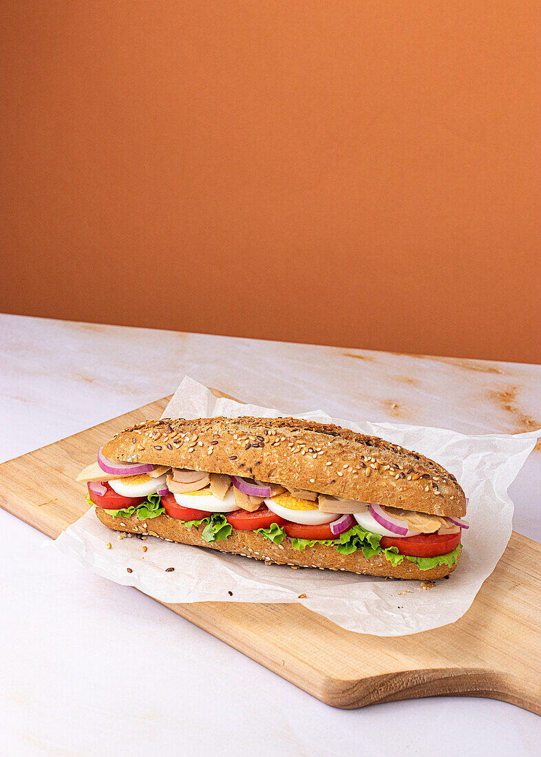 Appetizing sandwich with hard boiled egg, fresh lettuce, tuna and tomatoes served on baking paper on chopping wooden board on colorful background