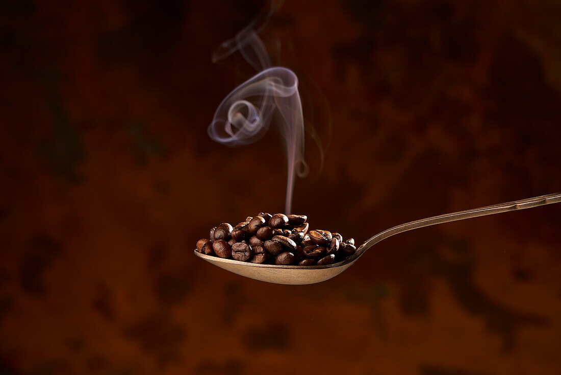 From above of closeup of spoon full of heap of roasted coffee beans on brown background